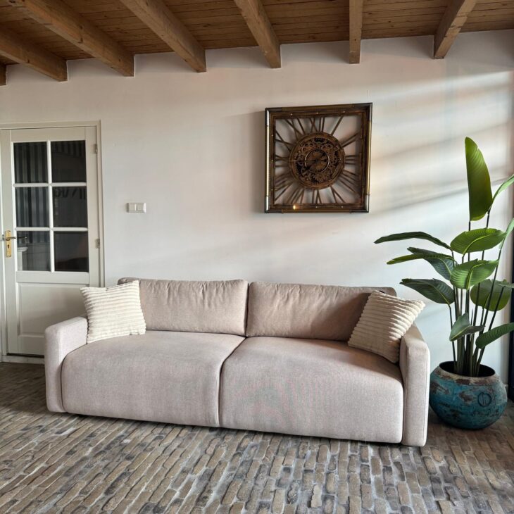 Beige tweezitsbank met losse kussens onder een grote wandklok