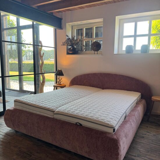 Oudroze gestoffeerd boxspringbed met rond hoofdbord onder een houten balkenplafond