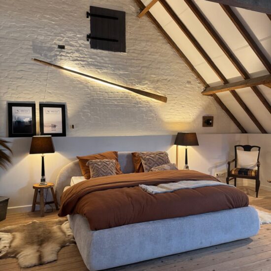 Beige gestoffeerd boxspringbed met roestbruin dekbed op een zolder met houten spanten