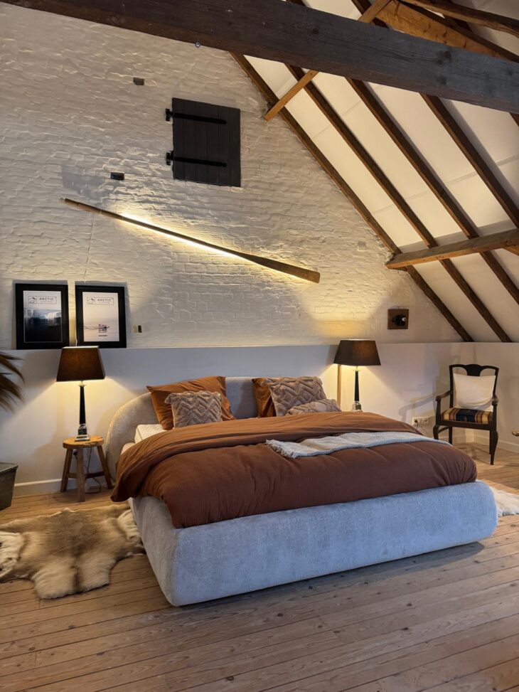 Beige gestoffeerd boxspringbed met roestbruin dekbed op een zolder met houten spanten